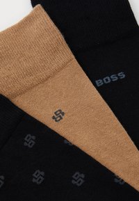 Trois paires de chaussettes sont exposées : une noire avec un logo sombre, une beige avec un logo similaire, et une paire noire texturée en haut.