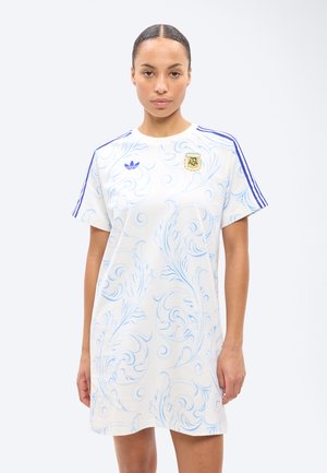 Frau trägt ein weißes Kleid, gestaltet wie ein Sporttrikot mit blauen Wirbeln, blauen Streifen auf den Schultern und einem AFA-Emblem auf der Brust.