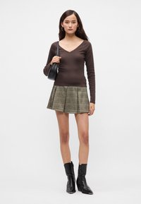 Καφέ μακριμάνικο μπλουζάκι με V-λαιμόκοψη συνδυασμένο με πλισέ πράσινη καρό φούστα. Μοντέλο που φορά μαύρες μπότες μέχρι τον αστράγαλο και κρατά μια μικρή μαύρη τσάντα.