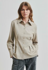 Lys beige skjorte med spiss krage, med plisserte sider og lange ermer, laget av glatt stoff.