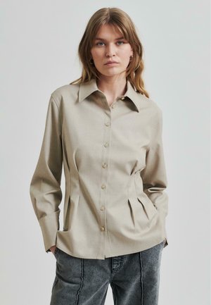 Lys beige skjorte med spiss krage, med plisserte sider og lange ermer, laget av glatt stoff.