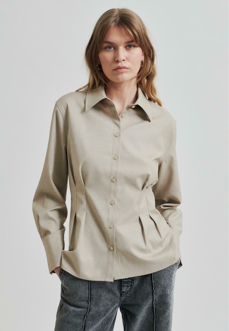 Lys beige skjorte med spiss krage, med plisserte sider og lange ermer, laget av glatt stoff.