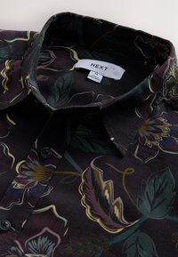 Chemise noire avec un motif floral dans des tons de violet, vert et jaune. Équipée d'un col à boutons et d'une étiquette indiquant la taille M.