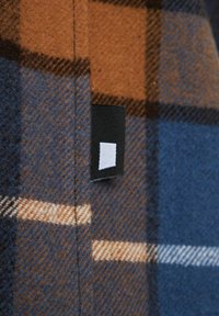 Brązowo-niebieska tkanina w kratę z białymi akcentami, o teksturowanym wykończeniu i czarną etykietą z białym kwadratowym detalem.