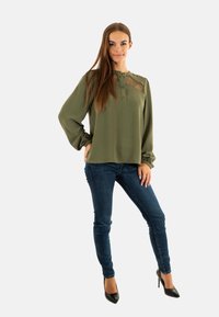 Blusa de manga larga verde oliva con cuello abotonado y acentos de encaje transparente, combinada con jeans ajustados azul oscuro y tacones negros.