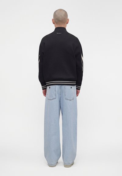 Personne avec la tête rasée portant une veste noire avec des bandes blanches sur les manches et un jean large bleu clair, vue de dos.