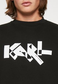 Czarna bluza z wyraźnym, geometrycznym białym logo "KARL" na przedzie, o gładkim, błyszczącym wykończeniu i klasycznym okrągłym dekolcie.