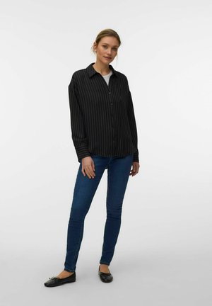 Kobieta stojąca, ubrana w czarną koszulę w prążki zapinaną na guziki, niebieskie obcisłe dżinsy i czarne baletki, na białym tle.