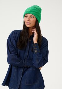 Zelena pletena kapa z rebrastim vzorcem in logotipom, v kombinaciji s temno modro jeans srajco z gumbnimi detajli in sprednjimi žepi.