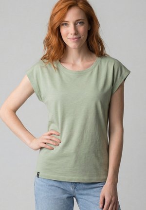 Junge Frau mit roten Haaren, die ein hellgrünes ärmelloses Oberteil und hellblaue Jeans trägt, steht mit einer Hand in der Hüfte.