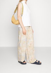 Kvinna i ärmlös vit topp, lösa pastellfärgade blommiga byxor, marinblå sandaler och bär en senapsgul halvmåneformad axelväska.