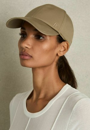 Beige Kappe mit einem runden Schirm und einem gestickten Logo an der Seite, getragen über einem weißen, strukturierten Oberteil mit Rundhalsausschnitt.