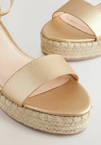 Zlatne espadrile imaju široke teksturirane trake, glatku unutrašnjost i platformu od pletenog jutea.