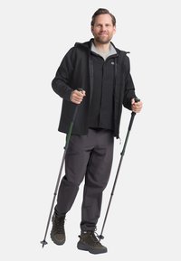 Schwarze wasserdichte Jacke mit einer grau gefütterten Kapuze, schwarzes Hemd darunter, dunkelgraue Hose und braune Wanderstiefel. Hält Trekkingstöcke.
