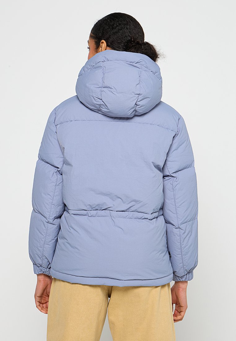 Persona che indossa una giacca a vento leggera di colore blu chiaro con un grande cappuccio, vista da dietro, abbinata a pantaloni beige su uno sfondo neutro.