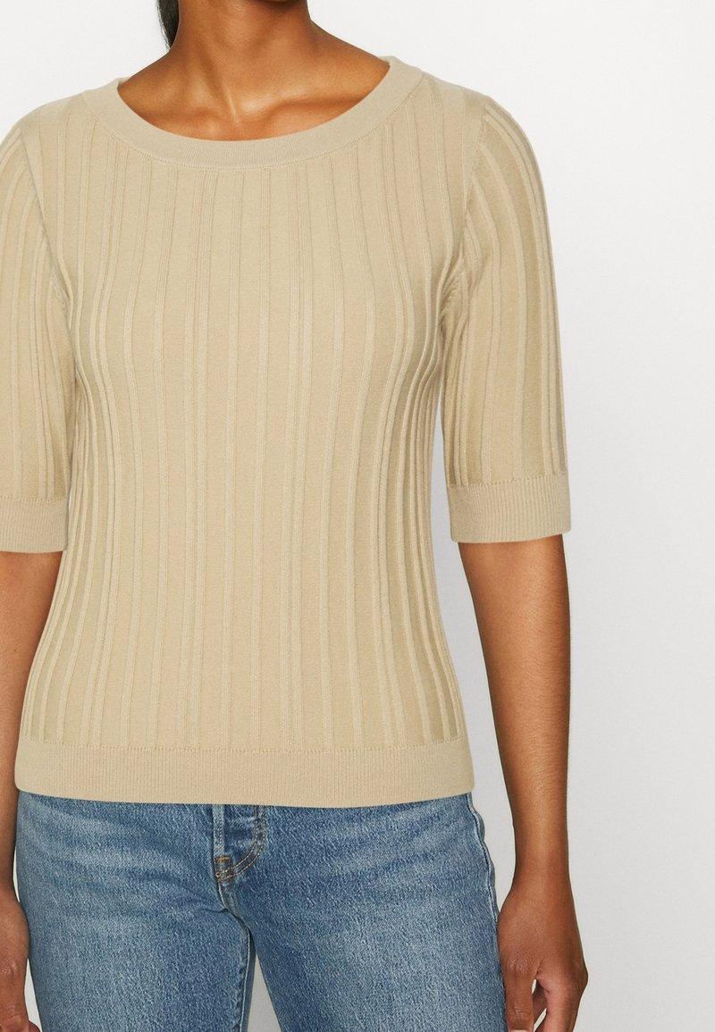 Suéter de punto acanalado beige con escote redondo y mangas cortas, que presenta una cintura ajustada y una textura suave. Combinado con jeans azules.