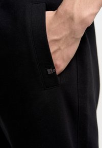 Zwarte sportbroek met een gladde textuur, voorzien van een zijzak en een klein zwart label met de tekst "BIG STAR" zichtbaar aan de rand van de zak.
