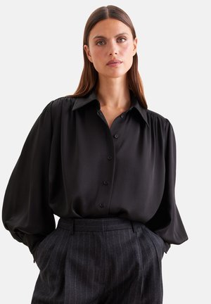Donna con lunghi capelli castani che indossa una camicetta nera ampia con bottoni e pantaloni a vita alta scuri a righe sottili, con le mani nelle tasche.