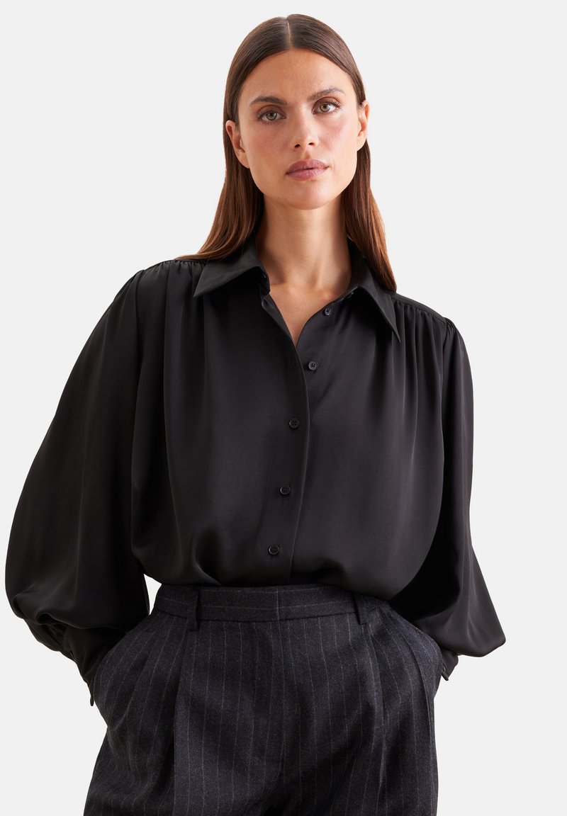Femme aux longs cheveux bruns portant une blouse noire ample à boutons et un pantalon à fines rayures foncé taille haute, les mains dans les poches.