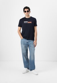 Mørkeblå bomulls T-skjorte med grafisk tekstdesign i rødt og hvitt. Kombinert med lyseblå jeans og hvite joggesko. Modell som bærer solbriller.