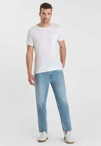 Camiseta blanca de algodón con un bolsillo, combinada con unos vaqueros de mezclilla azul claro. El modelo lleva zapatillas blancas con suela beige. Diseño sencillo.