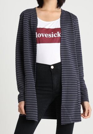 Vrouw met een open cardigan in blauw en zwart gestreept, een wit shirt met de tekst "lovesick" in een rood vak en een zwart broek met hoge taille.