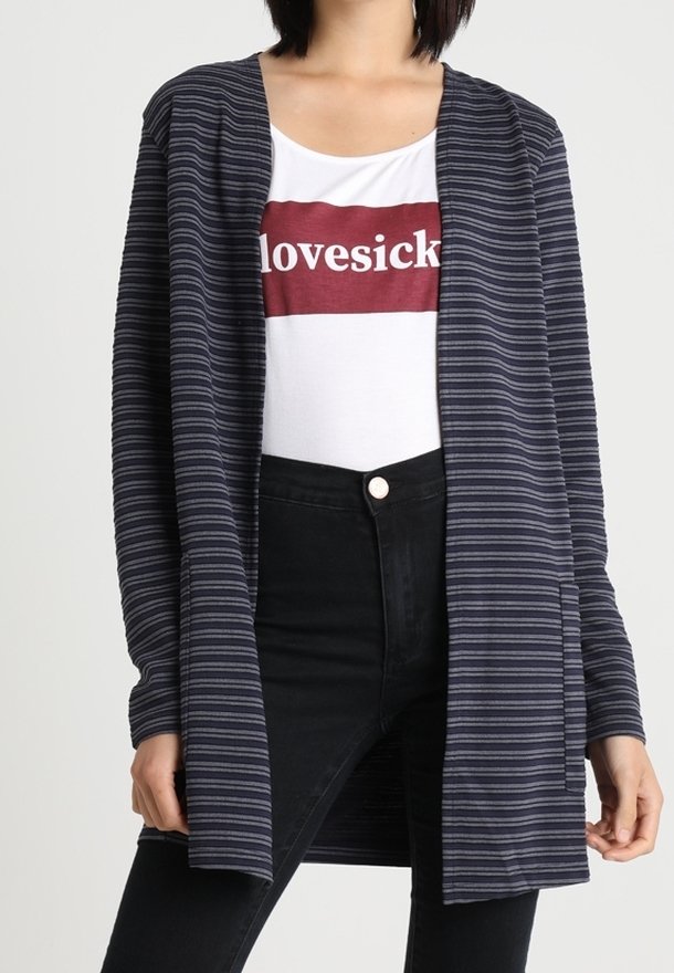 Femme portant un cardigan rayé bleu et noir ouvert, un chemisier blanc avec le texte "lovesick" dans un encadré rouge, et un jean noir taille haute.