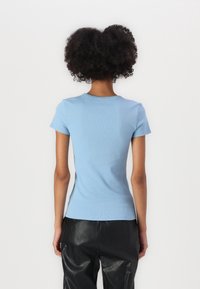 Osoba z kręconymi włosami, ubrana w dopasowaną jasnoniebieską koszulę z krótkim rękawem i czarne skórzane spodnie, odwrócona plecami na jednolitym białym tle.