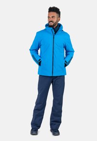 Chaqueta impermeable azul con capucha, con cierre de cremallera negra y bolsillos laterales. Combinada con pantalones azul marino, todo en una textura de tela lisa.