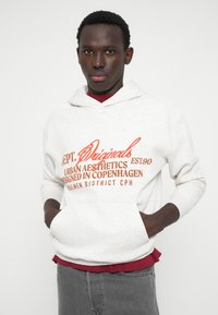 Hoodie cinza claro com bolso frontal, apresentando texto gráfico em vermelho-laranja. Material em mistura de algodão, capuz com cordão, sobre uma camisa vermelha.