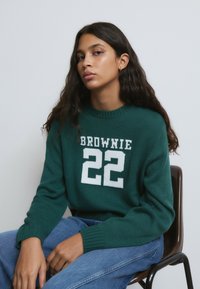 Suéter verde de punto con el texto blanco "BROWNIE 22". Puños y cuello acanalados. Modelo sentado en una silla, con jeans de denim azul.