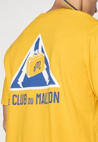Rumena bombažna majica s kratkimi rokavi, ki ima modro-belo grafiko. Na hrbtni strani je emblem s aktovko in besedilom "LE CLUB DU MALON."
