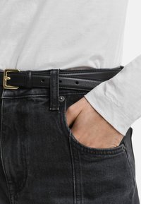 Hand in de zak van zwarte denim jeans met een zwarte leren riem, en de manchetten van een wit overhemd met lange mouwen zichtbaar.
