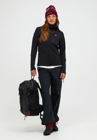 Black zip-up jacket, black pants, red hiking boots. Carrying a black backpack with red accents. Burgundy beanie with mountain graphics.