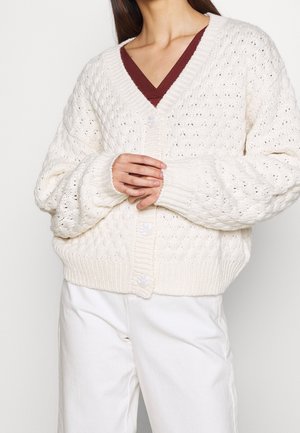 Frau trägt eine weiße grobgestrickte Strickjacke mit großen Knöpfen über einem bordeauxfarbenen V-Ausschnitt-Top und weißen Hosen, die Hände vor dem Körper verschränkt.