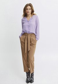 Lavendelfarbene Bluse mit langen Ärmeln und Kragen, in hochgeschnittene, braune gefältete Hose mit einem gebundenen Gürtel gesteckt, kombiniert mit schwarzen Cowboystiefeln.