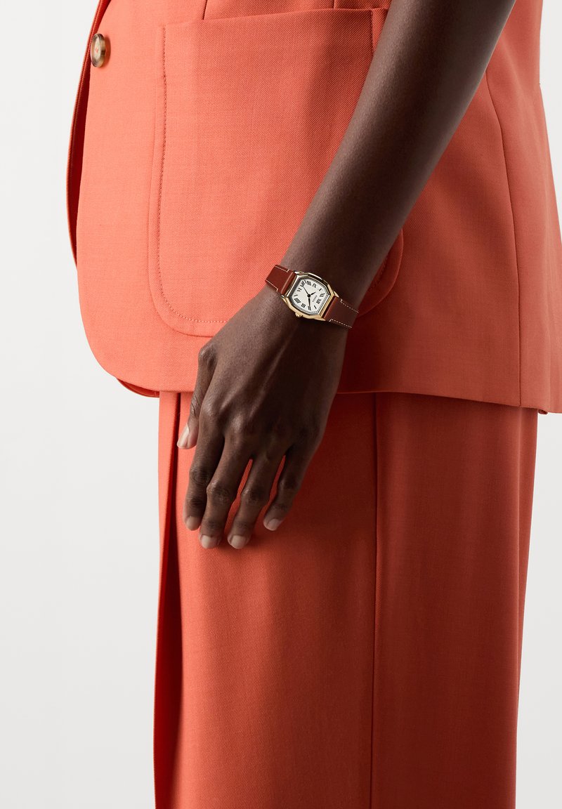 Montre-bracelet avec boîtier octogonal doré et bracelet en cuir marron portée au poignet gauche d'une personne en costume orange avec une grande poche avant.
