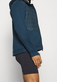 Veste à capuche en bleu sarcelle foncé avec un panneau avant matelassé, fermeture éclair et manches longues, associée à un short foncé. Texture du tissu lisse.