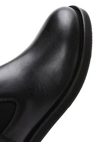 Primo piano della punta di un stivale Chelsea in pelle nera con finitura liscia, pannello elastico laterale e bordo della suola in gomma su sfondo bianco.