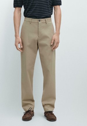 Mężczyzna ubrany w beżowe spodnie o prostych nogawkach z zapięciem na guzik i zamek, w połączeniu z brązowymi sznurowanymi butami oraz ciemną koszulą w paski z krótkim rękawem.