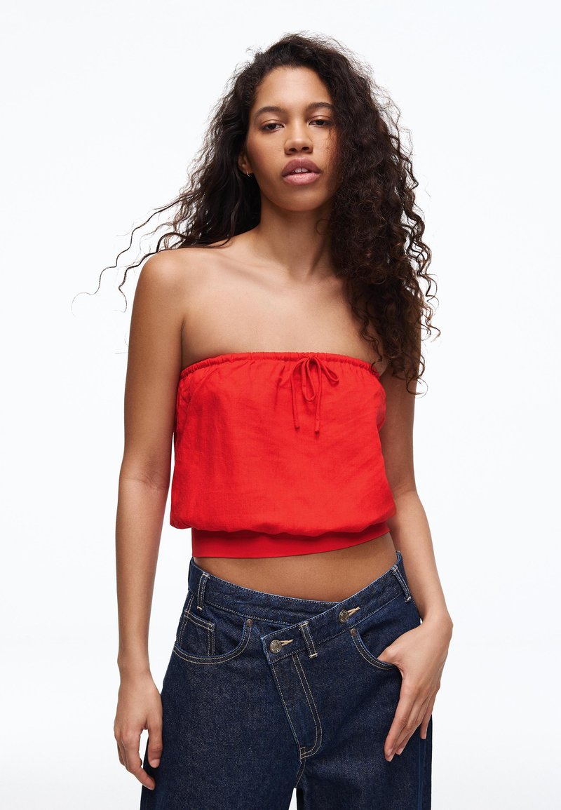 Jeune femme aux longs cheveux bouclés portant un haut rouge sans bretelles et un jean bleu foncé ample, posant devant un fond blanc.