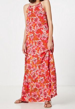 Kobieta ubrana w jasnoczerwoną suknię maxi bez rękawów z różowym i białym kwiatowym wzorem oraz brązowe płaskie sandały, stojąca na białym tle.