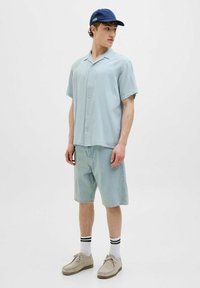 Camisa de botones de manga corta azul claro con un patrón sutil, combinada con pantalones cortos de mezclilla claros, zapatos grises y una gorra azul oscuro.