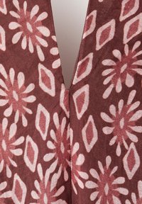 Zbliżenie na tkaninę w kolorze rdzy z jasnoróżowymi wzorami kwiatowymi i rombami oraz centralnym pionowym szwem lub otworem.