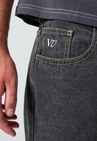 Μαύρο τζιν από τζιν με κίτρινα ραφές, με ροζ λογότυπο στην πίσω τσέπη. Το χέρι ακουμπάει στο πλάι του τζιν.