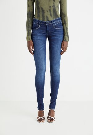 Jean slim en denim bleu foncé, avec une taille mi-haute, un design classique à cinq poches et des coutures contrastées. Assorti avec des sandales blanches à lanières.