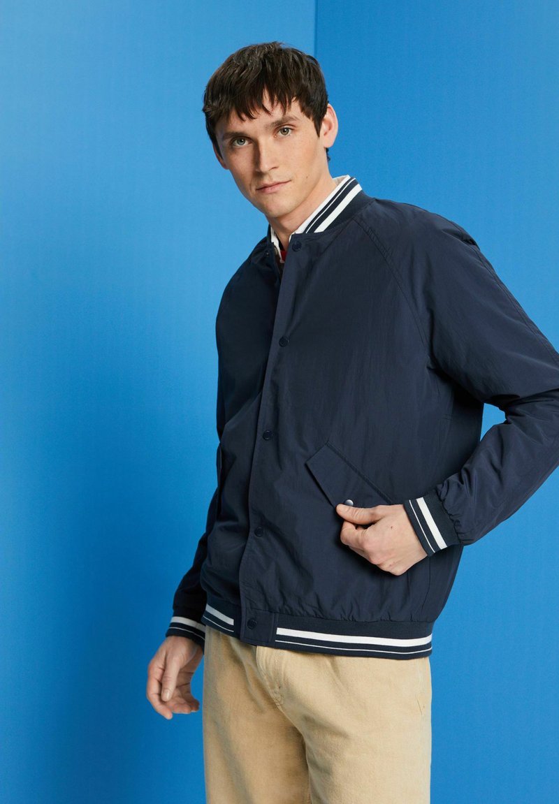 Jeune homme portant une veste de varsity bleu marine avec des rayures blanches et un pantalon beige, se tenant contre un fond bleu, la main dans la poche.