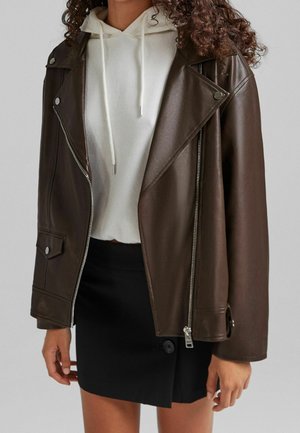 Femme portant une veste biker en cuir marron foncé, un sweat à capuche blanc et une mini-jupe noire à boutons sur fond uni.