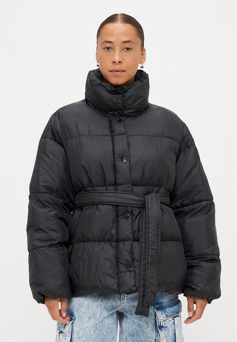 Casaco puffer preto com gola alta, fechos de botão, cintura com cinto e design matelassé. Apresenta uma textura suave e brilhante.
