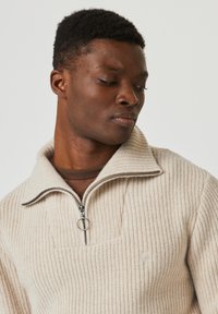 Pull à côtes crème avec un col zippé mi-long et une finition texturée subtile. Présente un tirette de zip ronde et un haut marron superposé en dessous.